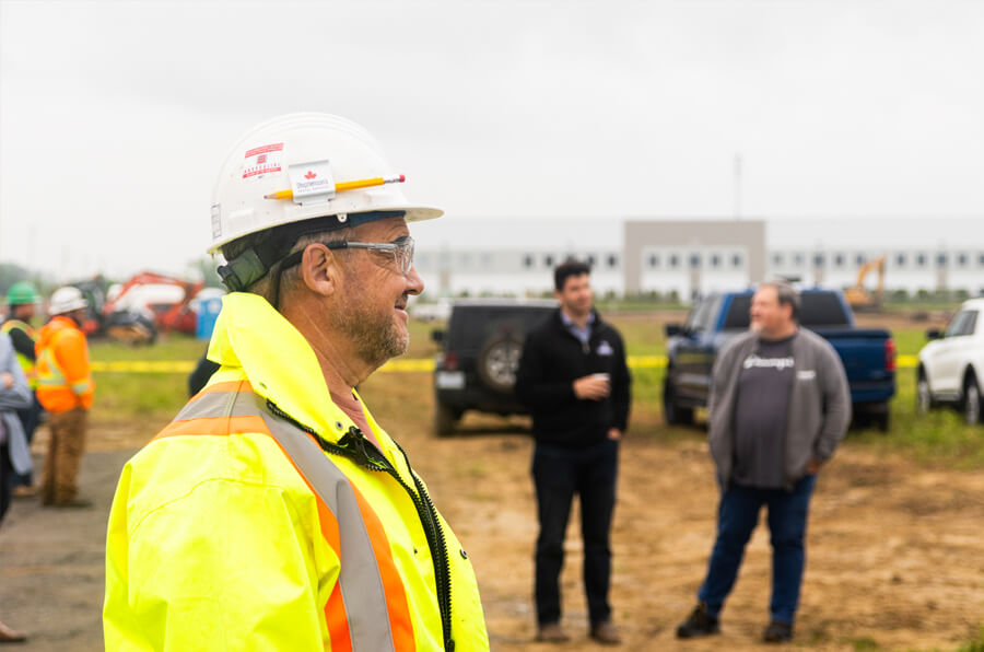 A construction worker in a yellow safety jacket and white helmet stands in the foreground, smiling. In the background, people are chatting near vehicles at a window production plant in Ottawa, ON.
